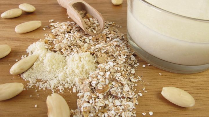 Haferflocken und Mandeln sind auf einem Holzbrett verstreut, w&auml;hrend ein Glas mit milchiger Fl&uuml;ssigkeit daneben steht.