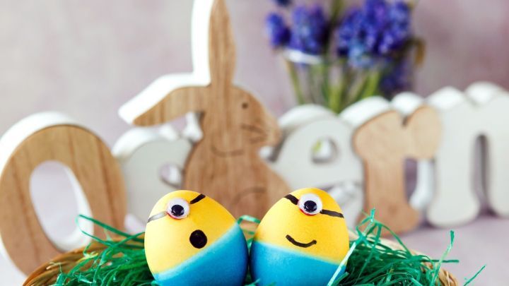 Bunte Ostereier im Minion-Design liegen in einem Nest aus Gras, umgeben von osterlichen Dekorationen und Blumen.