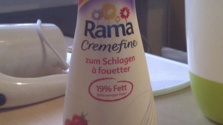 Eine Flasche Rama Cremefine steht auf einem Tisch, umgeben von K&uuml;chenutensilien. Sie enth&auml;lt fettarme Schlagsahne mit Erdbeermotiven.