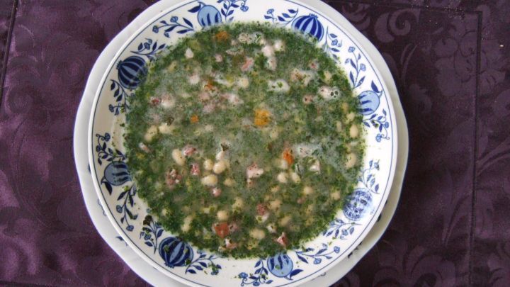 Eine Sch&uuml;ssel mit schneller wei&szlig;er Bohnensuppe ist gef&uuml;llt mit einer gr&uuml;nen, dicken Br&uuml;he und bunten Zutaten, auf einem lila Tischdeck.