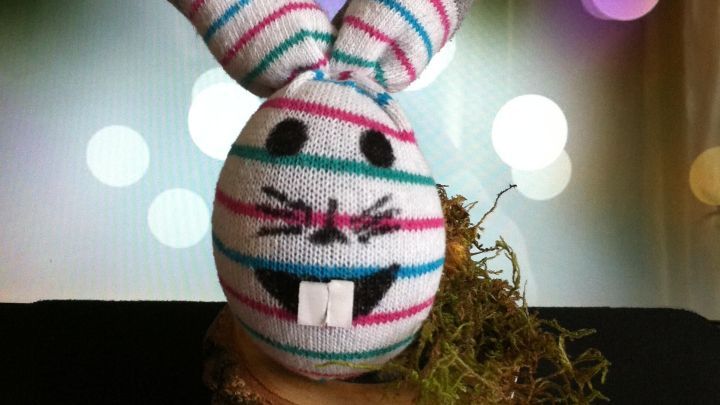 Ein lustiger Sockenhase mit gestreiften Ohren sitzt auf einem Holzscheit, umgeben von buntem, verschwommenem Licht.