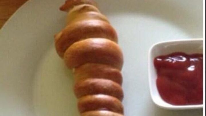 Ein warmes W&uuml;rstchen im Brezelmantel liegt auf einem Teller mit einer Schale Ketchup, umgeben von einem bunten Salat.