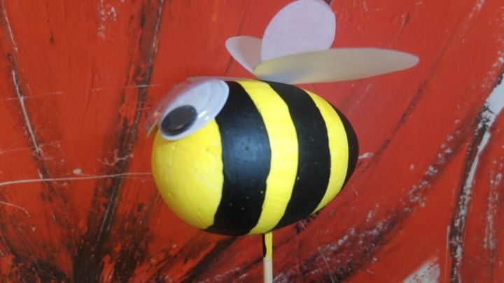 Eine gelb-schwarze Bastel-Hummel mit Fl&uuml;geln steht auf einem Stock vor einer roten, blumigen Hintergrundgestaltung.