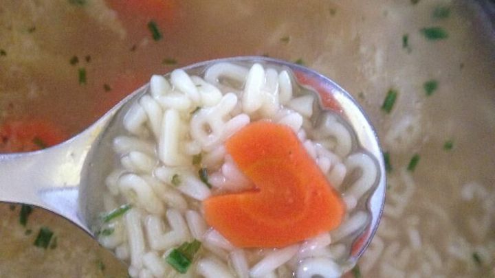 Eine L&ouml;ffel mit Herzchen-karotte hebt sich &uuml;ber eine Suppe mit Nudeln und frischen Kr&auml;utern.