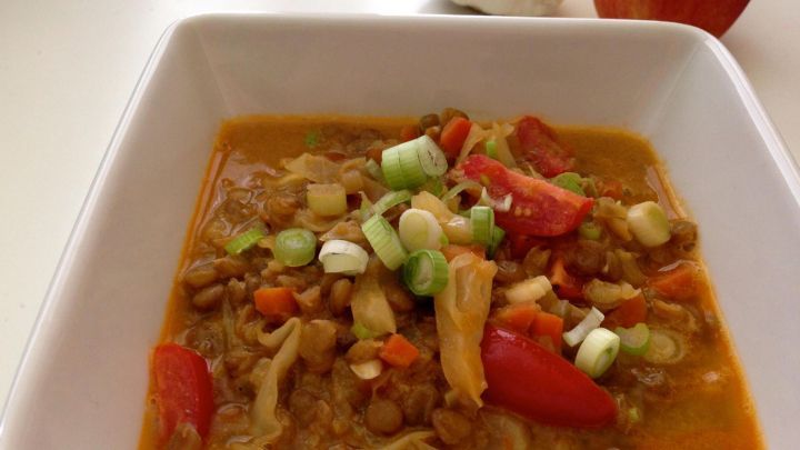 Ein veganer Linsen-Kokos-Kohl Eintopf in einer Sch&uuml;ssel, garniert mit frischem Gem&uuml;se, auf einem hellen Tisch.