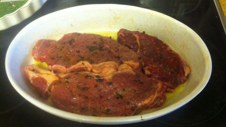 Rindersteaks ruhen in Oliven&ouml;l und Pfeffer in einer ovalen, wei&szlig;en Auflaufform auf einem dunklen Herd.