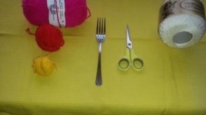 Bunte Wolle und eine Schere liegen auf einem gelben Tuch, w&auml;hrend eine Gabel zur Herstellung von Bommeln bereitsteht.