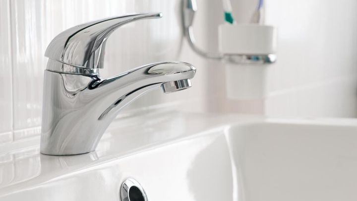 Ein gl&auml;nzender Wasserhahn flie&szlig;t &uuml;ber ein wei&szlig;es Waschbecken in einem hellen Badezimmer mit Zahnb&uuml;rsten im Hintergrund.