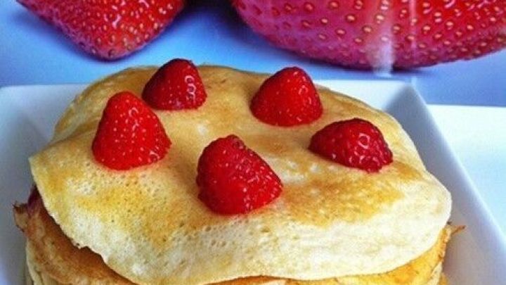 Gestapelte, goldbraune Pfannkuchen sind mit frischen Erdbeeren bedeckt, im Hintergrund sind gro&szlig;e Erdbeeren zu sehen.