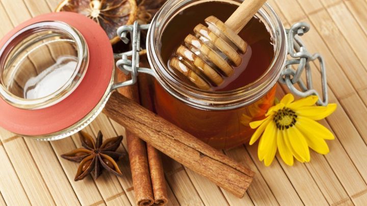 Ein Glas Honig mit einem Holzl&ouml;ffel steht auf einer Bambusmatte, umgeben von Zimtstangen und bl&uuml;henden Pflanzen, die das Immunsystem unterst&uuml;tzen.