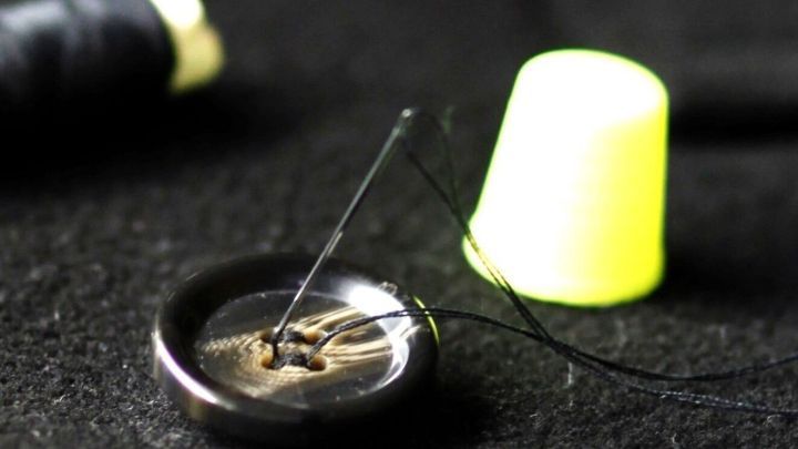 Ein schwarzer Knopf liegt auf dunklem Untergrund. Eine Nadel mit schwarzem Faden ist eingestochen. Ein gelber Fingerhut steht daneben.