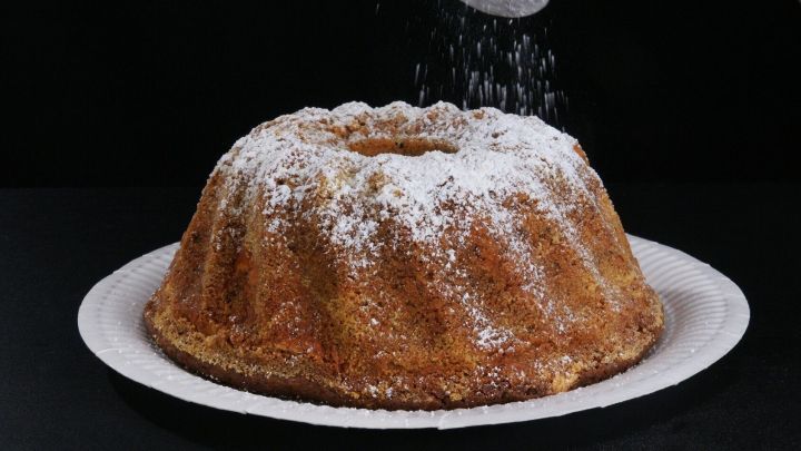 Ein Sandkuchen mit Amarettini und Schokolade wird mit Puderzucker best&auml;ubt, pr&auml;sentiert auf einem wei&szlig;en Teller vor schwarzem Hintergrund.