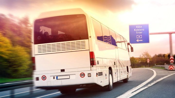 Ein Reisebus f&auml;hrt schnell auf einer Autobahn, umgeben von gr&uuml;nen B&auml;umen und unter einem bunten Himmel.