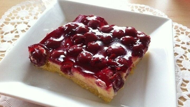 Ein St&uuml;ck Blechkuchen mit Kirschen und gl&auml;nzender Vanillepudding-F&uuml;llung liegt auf einem wei&szlig;en Teller mit Spitze.