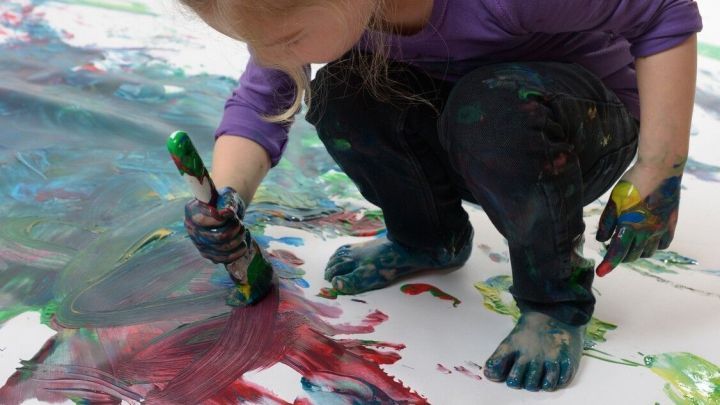 Ein kleines M&auml;dchen mit blauen H&auml;nden malt mit Fingerfarben auf einem gro&szlig;en wei&szlig;en Blatt auf dem Boden.