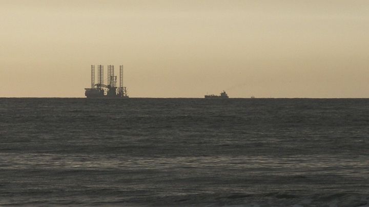 Eine Bohrinsel steht stabil im Wasser, w&auml;hrend ein kleines Schiff vorbeif&auml;hrt, unter einem sanften Sonnenuntergang.