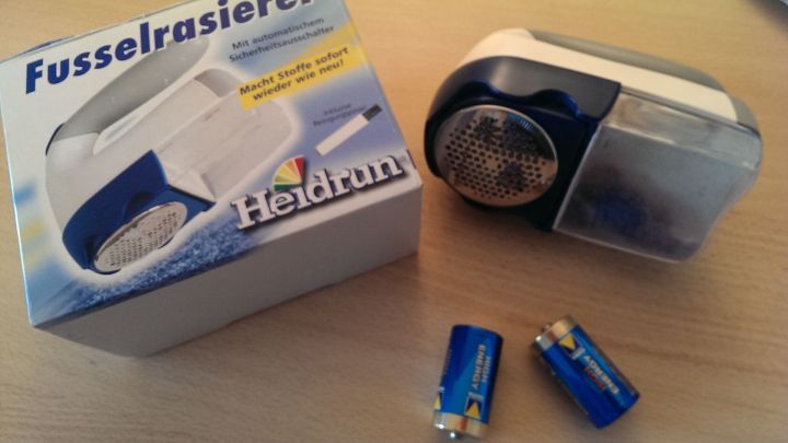Ein Fusselrasierer von Heidrun liegt neben seiner Verpackung und zwei Batterien auf einem Tisch und ist bereit zur Nutzung.