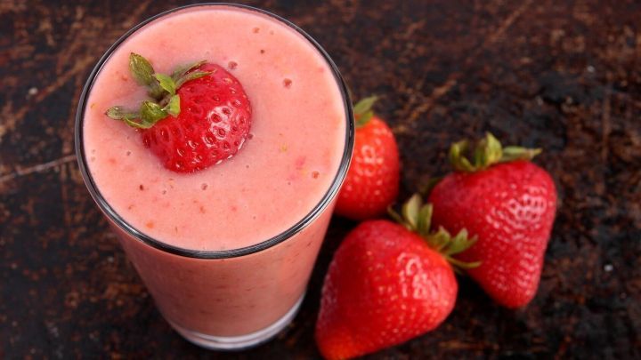 Ein Erdbeer-Smoothie in einem Glas mit einer Erdbeere dekoriert, umgeben von frischen Erdbeeren auf einem dunklen Tisch.