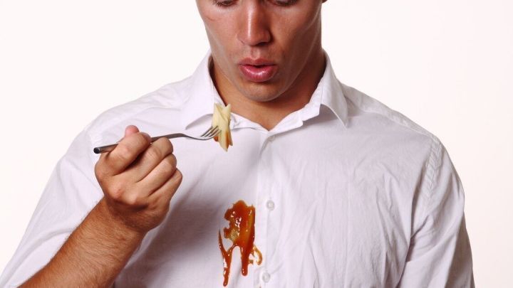 Ein Mann mit einem wei&szlig;en Hemd hat So&szlig;e auf seiner Kleidung. Er schaut &uuml;berrascht auf einen L&ouml;ffel Nudeln.