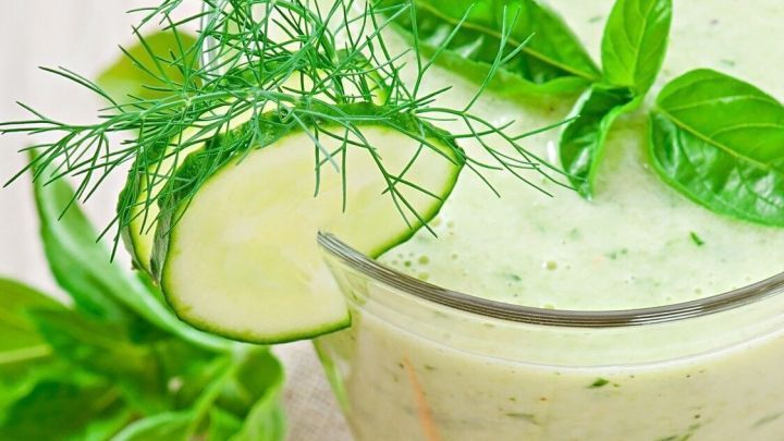 Ein Gurkensmoothie steht auf einem Tisch, garniert mit Basilikum und Dill, daneben liegen Chilis und ein Brotst&uuml;ck.