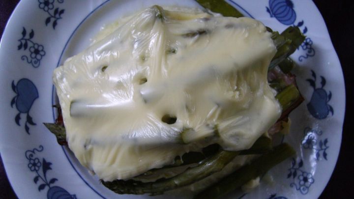 &Uuml;berbackener gr&uuml;ner Spargel liegt auf einem dekorativen Teller und wird von einer cremigen K&auml;seschicht &uuml;berzogen.