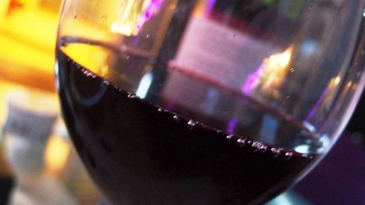 Ein rot gef&uuml;lltes Weinglas steht auf einem Tisch, umgeben von unscharfen bunten Lichtern, die f&uuml;r eine gem&uuml;tliche Atmosph&auml;re sorgen.