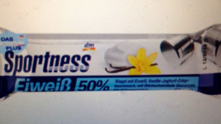 Ein Proteinriegel liegt auf einem hellen Hintergrund, beschriftet mit &bdquo;Sportness Eiwei&szlig; 50%&ldquo; und einer Vanilleillustration.