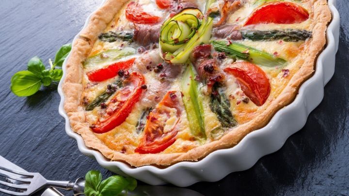 Eine frisch gebackene Quiche mit gr&uuml;nem Spargel und Tomaten liegt dekorativ in einer wei&szlig;en Tarteform auf einem dunklen Tisch.