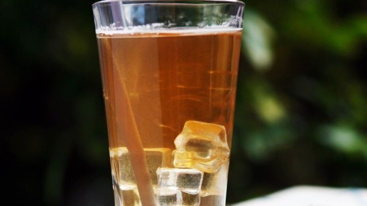 Ein Glas mit Eistee und Eisw&uuml;rfeln steht auf einem Tisch im Freien, umgeben von gr&uuml;ner Pflanzenwelt.