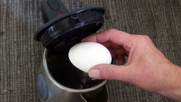 Ein Hand greift ein rohes Ei und legt es in einen ge&ouml;ffneten Wasserkocher, um Fr&uuml;hst&uuml;ckseier zuzubereiten.