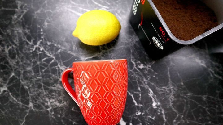 Eine rote Tasse liegt auf einem marmorierten Tisch neben einer Zitrone und einer Kaffeetasse mit gemahlenem Kaffee.