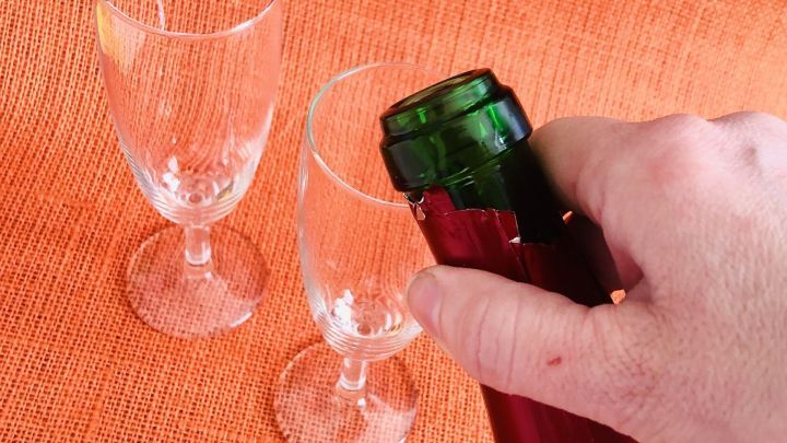 Eine Hand &ouml;ffnet eine Sektflasche vorsichtig &uuml;ber zwei Gl&auml;ser, um Schaum zu vermeiden, vor einem leuchtend orangefarbenen Hintergrund.