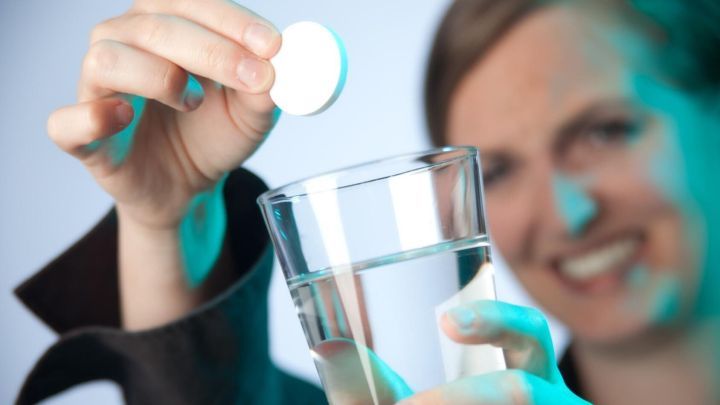 Eine Person h&auml;lt eine Brausetablette &uuml;ber ein Glas Wasser, w&auml;hrend sie l&auml;chelt und es in einer neutralen Umgebung betrachtet.