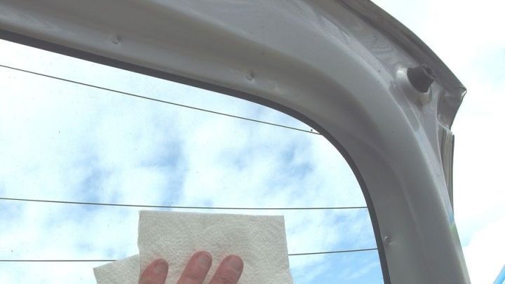 Eine Hand mit einem Tuch reinigt eine Autoscheibe, um Schlieren zu entfernen, w&auml;hrend blauer Himmel im Hintergrund sichtbar ist.