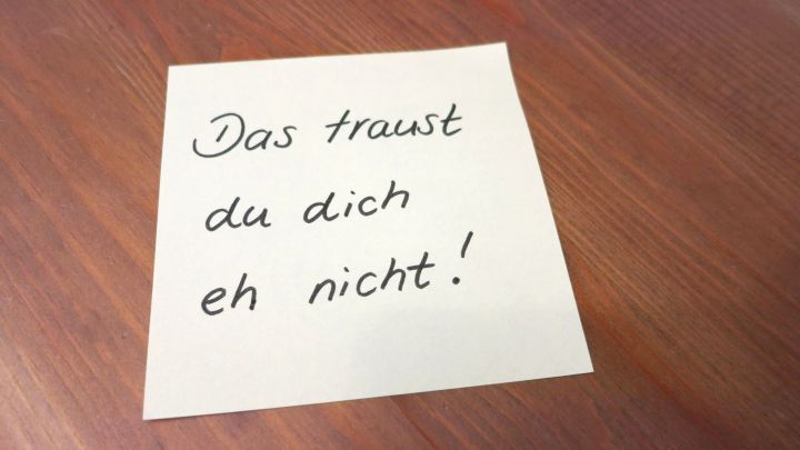 Ein gelbes Papier mit handgeschriebener Aufforderung liegt auf einem Holztisch und sagt: "Das traust du dich eh nicht!"
