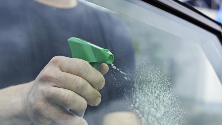 Eine Hand h&auml;lt eine Spr&uuml;hflasche, w&auml;hrend sie Scheiben mit Wasser bespr&uuml;ht, um die Autoscheibe zu reinigen.