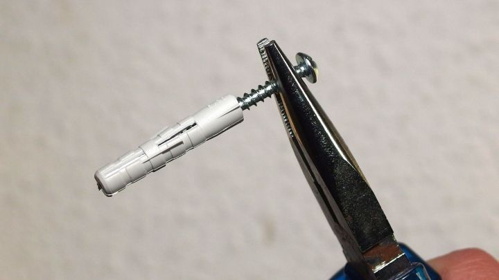 Eine Zange h&auml;lt eine Schraube, die in einen D&uuml;bel eingesetzt ist; im Hintergrund ist eine neutrale Wand zu sehen.