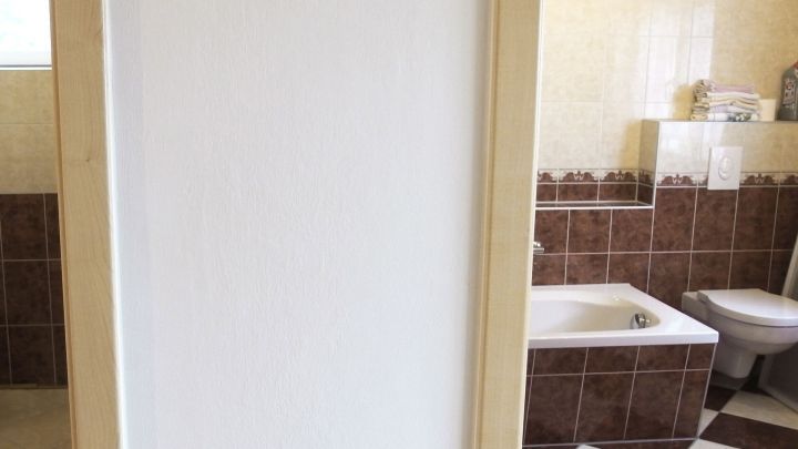 Eine wei&szlig;e Wand ist im Vordergrund, w&auml;hrend im Hintergrund ein Badezimmer mit braunen Fliesen und einer Badewanne sichtbar ist.