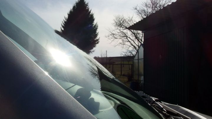 Ein Auto mit einer gewellten Windschutzscheibe steht hell erleuchtet im Freien, umgeben von B&auml;umen und einem Schuppen.