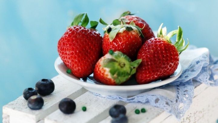 Rote Erdbeeren liegen auf einem wei&szlig;en Teller mit einem Spitzenserviette, umgeben von blauen Beeren und einer hellblauen Hintergrund.