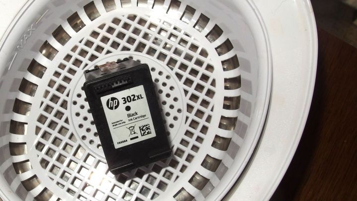 Ein HP 302XL Tintenpatrone liegt in einem wei&szlig;en Gitterkorb eines Ultraschallbads, bereit zur Reinigung.