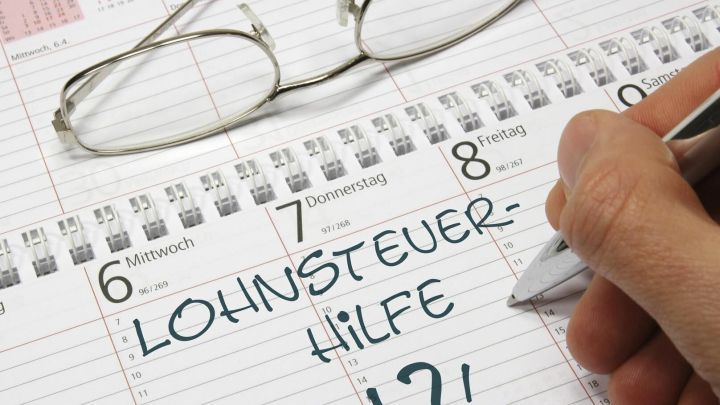 Auf einem Kalender schreibt eine Person mit einem Stift "LOHNSTEUER-HILFE" und schaut durch eine Lesebrille, um wichtige Termine zu notieren.