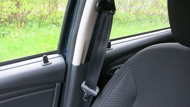 Ein Sicherheitsgurt im Auto h&auml;ngt ungenutzt an der R&uuml;ckseite des Fahrersitzes, w&auml;hrend im Hintergrund gr&uuml;ne B&auml;ume sichtbar sind.