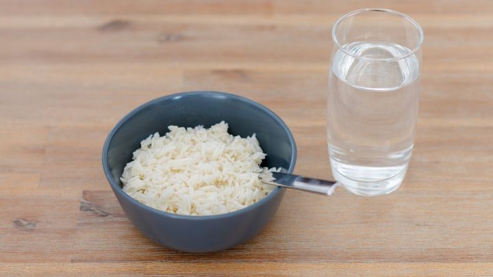 Eine Sch&uuml;ssel mit Reis und ein Glas Wasser stehen auf einem Holztisch; der L&ouml;ffel liegt in der Sch&uuml;ssel.