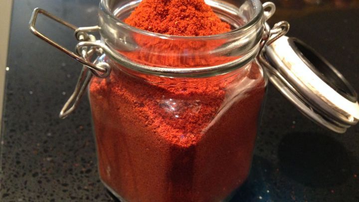 Ein Glas mit Paprikapulver ist ge&ouml;ffnet, wobei eine mound von rotem Gew&uuml;rz im Inneren sichtbar ist. Der gl&auml;nzende Untergrund bietet einen starken Kontrast zu den leuchtenden Farben.