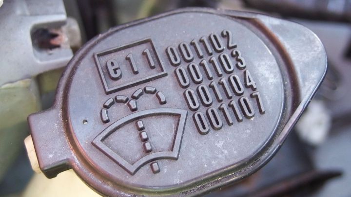 Ein schwarzes Beh&auml;lterdeckel zeigt Symbole und Zahlen f&uuml;r Scheibenwaschmittel, montiert im Motorraum eines Fahrzeugs.