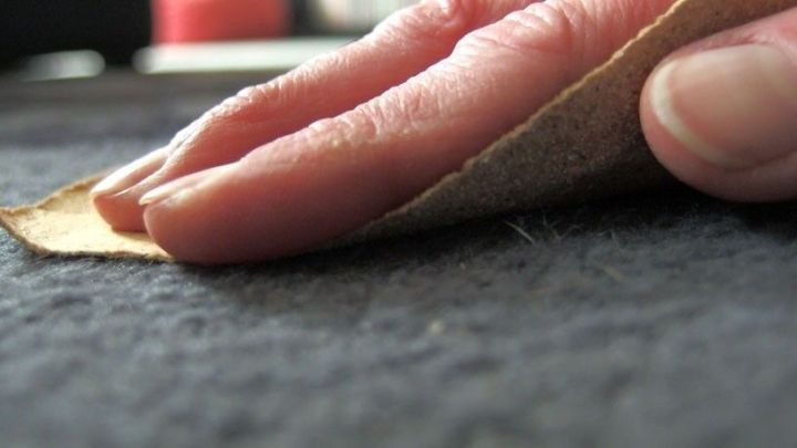Hand reibt mit Sandpapier &uuml;ber Teppich, um Tierhaare zu entfernen. Nahaufnahme zeigt Detailarbeit in h&auml;uslicher Umgebung.