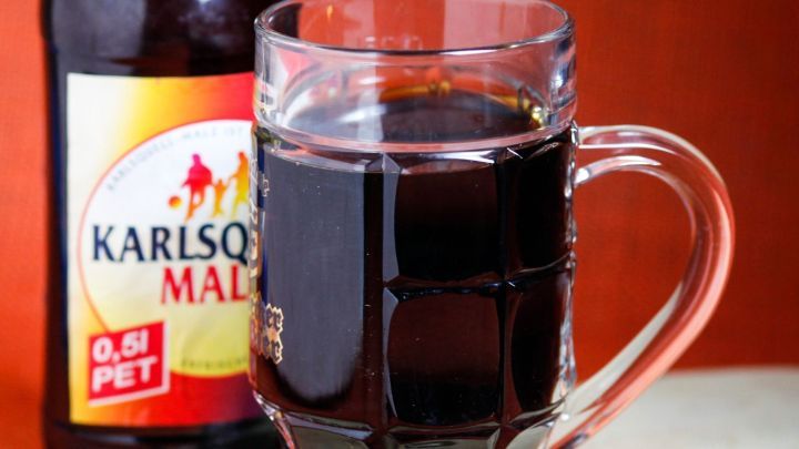 Ein Glas Malzbier steht auf einem Holzbrett, daneben eine Flasche mit dem Label "Karl&rsquo;s Malz". Der Hintergrund ist orange.