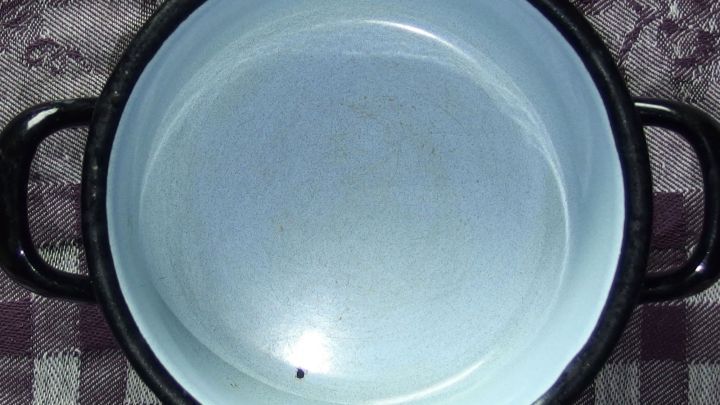 Ein Emailletopf liegt auf einem karierten Tisch, innen sichtbar sind Metallspuren, die auf Gebrauchsspuren hinweisen.