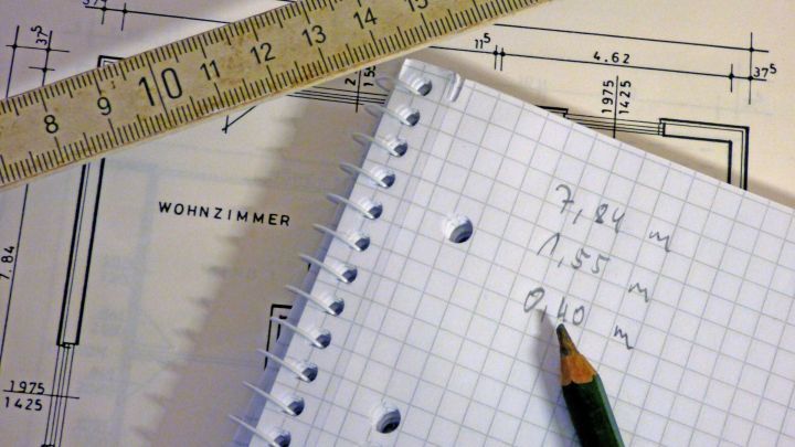 Ein Ma&szlig;band liegt &uuml;ber einem Geb&auml;udeplan mit Skizzen, w&auml;hrend ein Bleistift auf kariertem Papier f&uuml;r Wohnfl&auml;chenberechnungen verwendet wird.